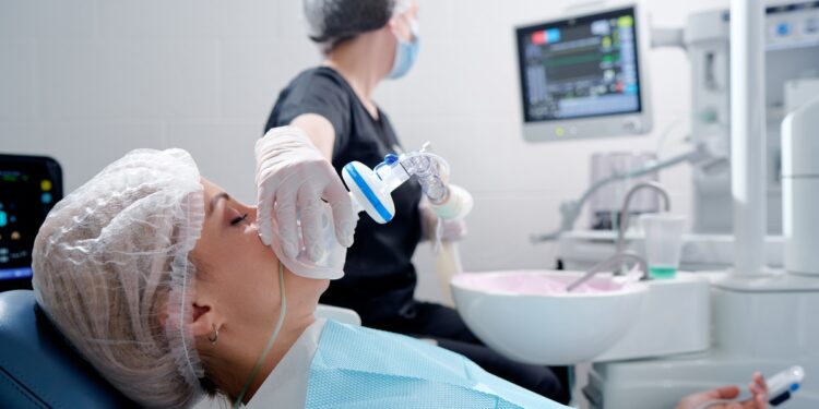 A woman lying in a dental chair while a gloved person holds an anesthesia mask near her face during dental care.
