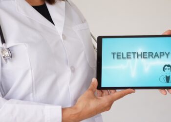 A close-up of a woman in a doctor's coat with a stethoscope holding an electronic tablet that says "TELETHERAPY."