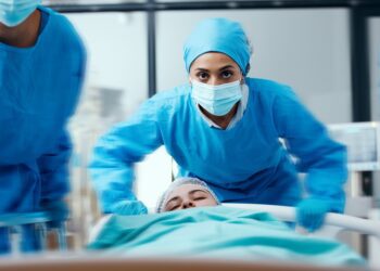 three people in blue surgical clothing and masks pushing a bed with a covered person in a bright hospital room.
