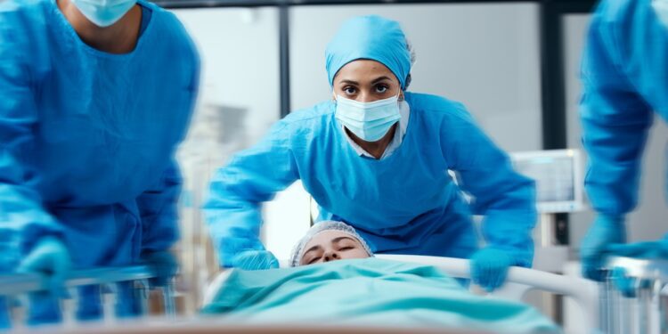 three people in blue surgical clothing and masks pushing a bed with a covered person in a bright hospital room.