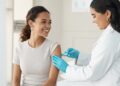 A woman in a white coat and blue gloves gives an injection to another woman sitting in a bright medical room.