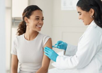 A woman in a white coat and blue gloves gives an injection to another woman sitting in a bright medical room.