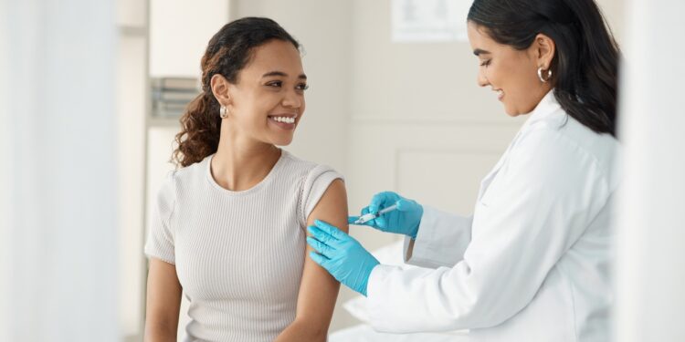 A woman in a white coat and blue gloves gives an injection to another woman sitting in a bright medical room.