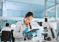 A woman in a white lab coat and latex gloves writes something down in a clipboard. A microscope is in front of her.
