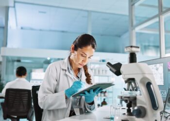 A woman in a white lab coat and latex gloves writes something down in a clipboard. A microscope is in front of her.
