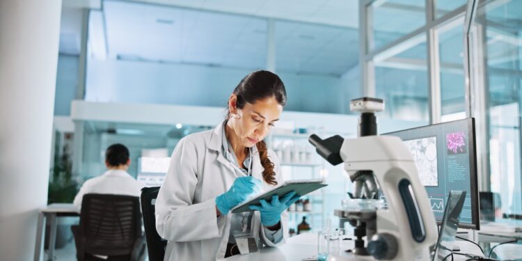 A woman in a white lab coat and latex gloves writes something down in a clipboard. A microscope is in front of her.
