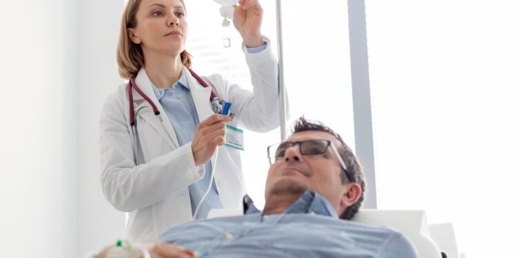 A man lies in a chair with an IV in his hand. A woman connects the IV tube to an IV bag behind him.