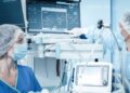A masked person in a blue uniform points at a monitor beside another masked person and medical equipment.