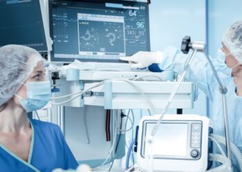 A masked person in a blue uniform points at a monitor beside another masked person and medical equipment.