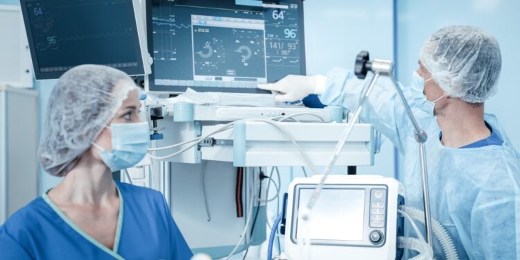 A masked person in a blue uniform points at a monitor beside another masked person and medical equipment.