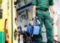 A person in a green uniform holding a blue medical bag while standing beside an open yellow ambulance.