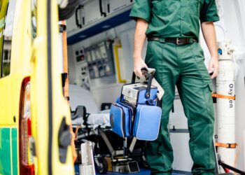 A person in a green uniform holding a blue medical bag while standing beside an open yellow ambulance.