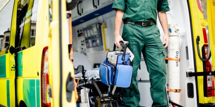 A person in a green uniform holding a blue medical bag while standing beside an open yellow ambulance.
