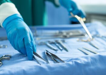 A person in a blue surgical gown and white gloves arranging metal instruments on a blue draped table.