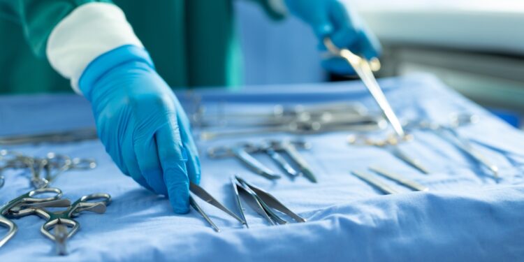 A person in a blue surgical gown and white gloves arranging metal instruments on a blue draped table.