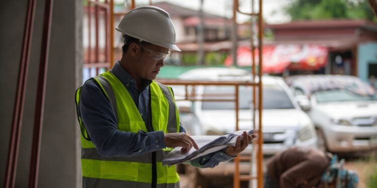 A worker in a white hard hat and yellow jacket holding large blueprints while standing near metal scaffolding.