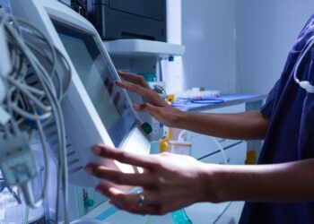 A person in blue scrubs touching a medical device with tubes, controls, and a screen in a hospital room.