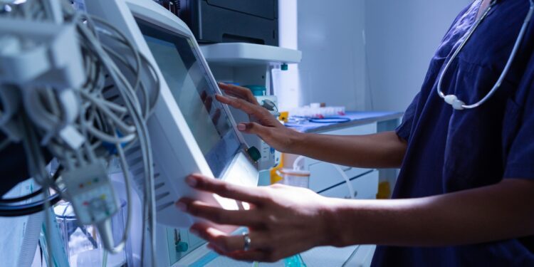 A person in blue scrubs touching a medical device with tubes, controls, and a screen in a hospital room.
