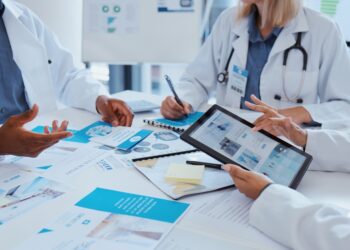 Three people in white coats meet at a table as one person points to a tablet above papers in a bright, medical office.
