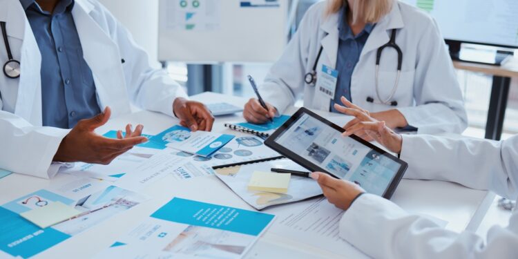 Three people in white coats meet at a table as one person points to a tablet above papers in a bright, medical office.