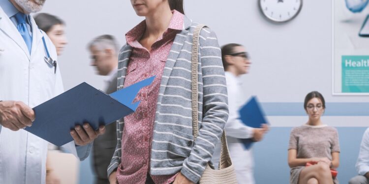 A man in a white coat holding a folder beside a woman in a bright hallway with seated people behind them.