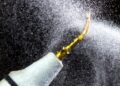 A white dental ultrasonic scaler with a curved gold tip is spraying water droplets against a black background.