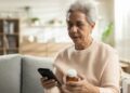 An aging adult woman sits on her couch at home to use her phone for ordering a prescription bottle refill.