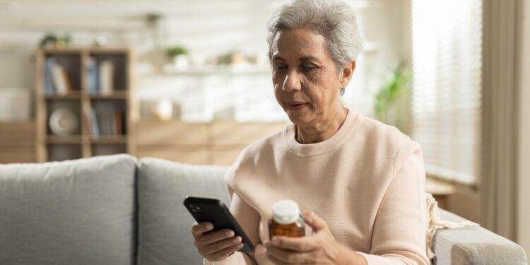 An aging adult woman sits on her couch at home to use her phone for ordering a prescription bottle refill.