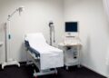 A room with black floors, a white curtain hanging from the ceiling bundled to the side, and a medical bed.