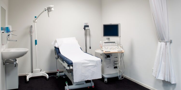 A room with black floors, a white curtain hanging from the ceiling bundled to the side, and a medical bed.