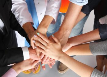 People in varied professional attire stack their hands together in the center, symbolizing teamwork and unity.