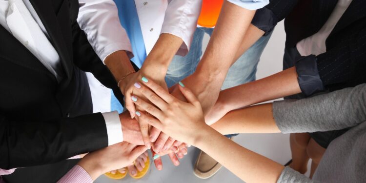 People in varied professional attire stack their hands together in the center, symbolizing teamwork and unity.