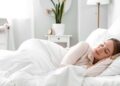 A woman is sleeping in a bed. She’s under some white covers and there’s a window on the wall.
