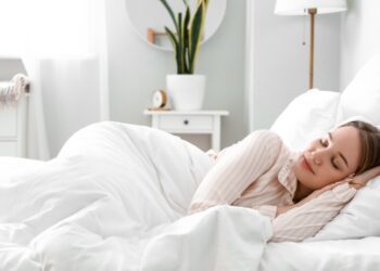 A woman is sleeping in a bed. She’s under some white covers and there’s a window on the wall.