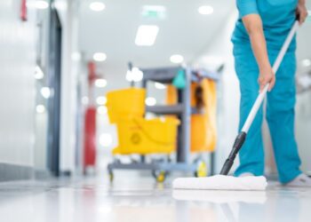 A person in blue scrubs mopping a shiny hospital hallway floor beside a yellow cleaning cart in the background.