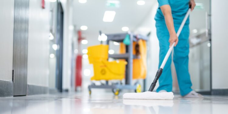 A person in blue scrubs mopping a shiny hospital hallway floor beside a yellow cleaning cart in the background.