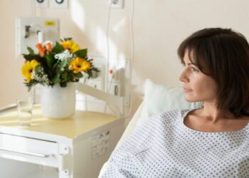 A woman with short brown hair wears a medical gown and lies in a hospital bed. She looks to the side toward a window.