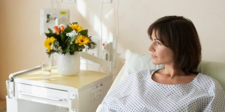 A woman with short brown hair wears a medical gown and lies in a hospital bed. She looks to the side toward a window.